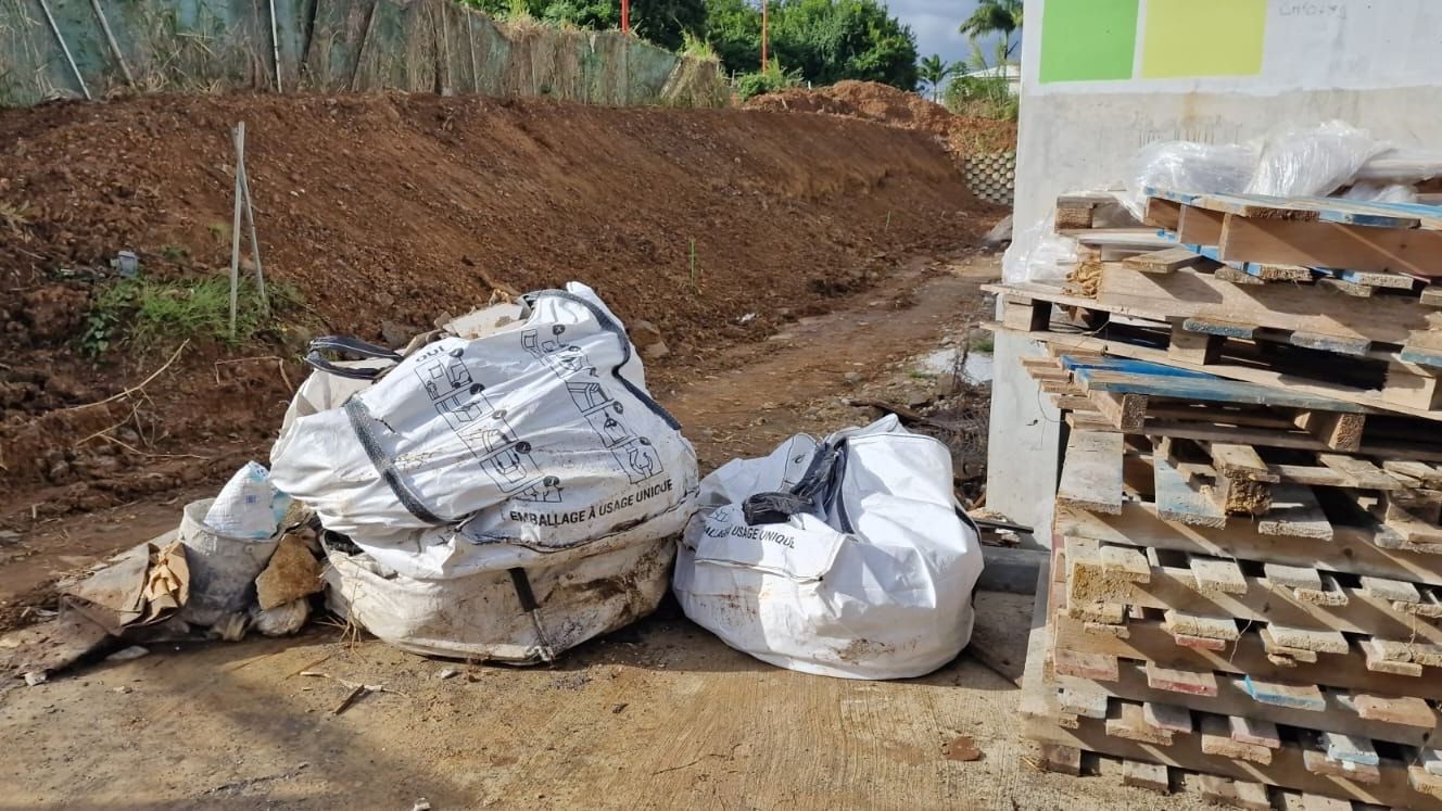 Deux big bags remplis de déchets posés au pied d'un talus de terre, à côté d'une pile de palettes